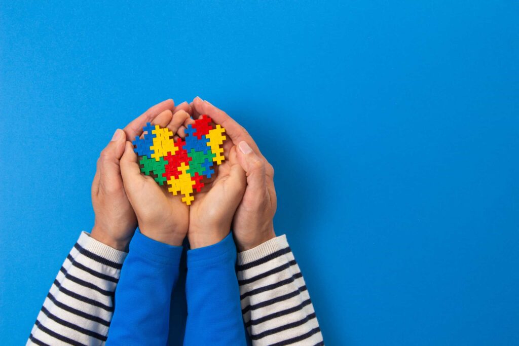 A photograph of a child and adults hands working together to hold a multi-coloured heart representing neurodiversity.
