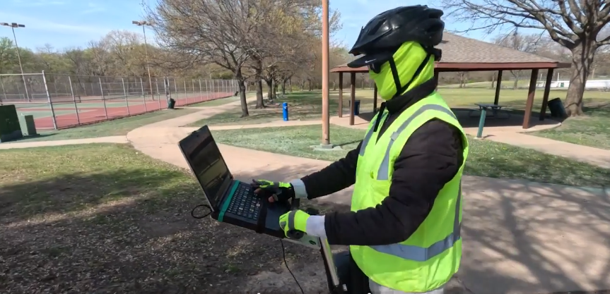 A man wearing a hi-vis jacket and sunglasses types on a laptop on a segway machine in an empty park in Texas, logging his findings.