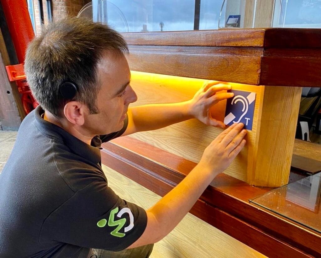 A man places a Hearing Loop sticker onto the underside of a desk.