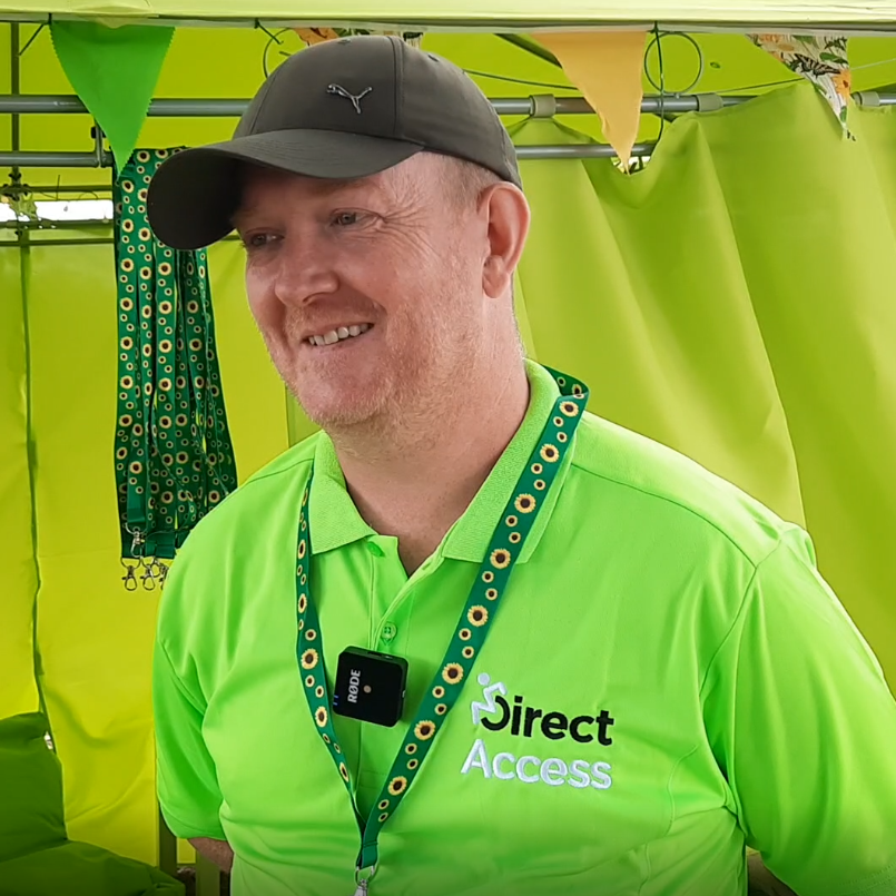 A photograph of Field Service Engineer Andy Lomas wearing a green Direct Access polo and cap in front of the Direct Access Calm Corner marquee. Sunflower lanyard can be seen hanging from the entrance of the tent.