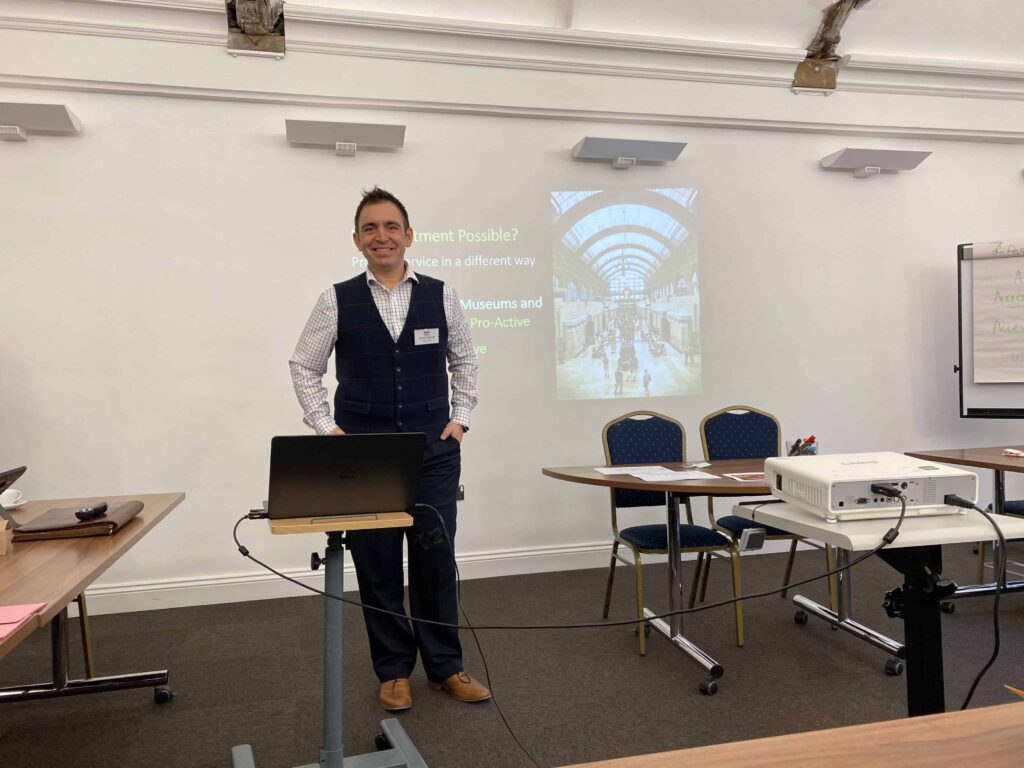 A photograph of a man, Steven Mifsud, delivering a talk about accessibility inclusion training, a powerpoint projected on the wall behind him. In front of him is a stand with his laptop.