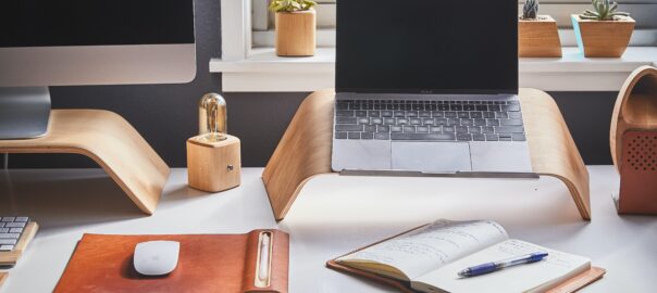 A person's workstation featuring a Mac computer, small laptop on a wooden stand, and an open notepad with a ballpoint pen sat atop it. In the background, a windowsill with a modest collection of cactus plants.