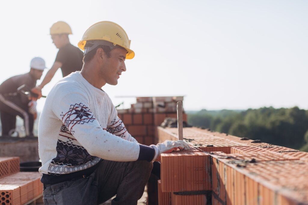 A male construction workers measures the level of a building being constructed as his colleagues move bricks in the background.
