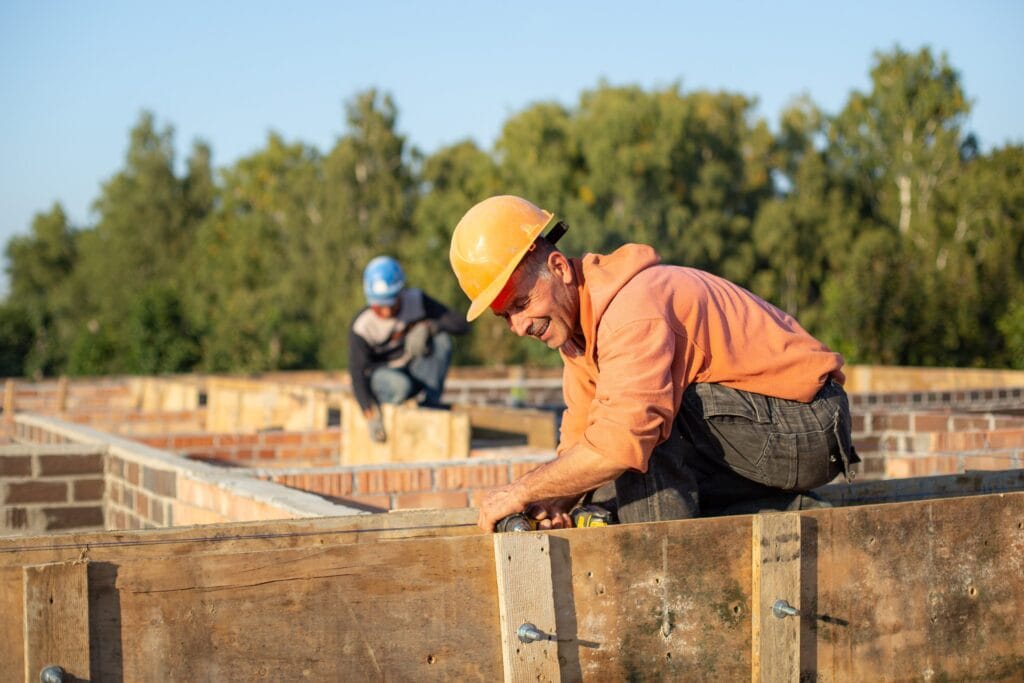 A man working on construction of the building drills into a piece of wood as a colleague works in the background moving a plank of wood.