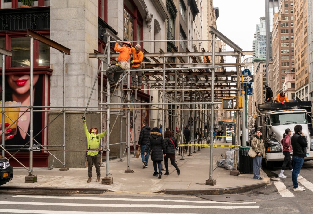 A street view of male builders on scaffolds working on construction of a building on the corner of a New York street. A man can be seen lifting a part of the scaffold for a colleague above as a young woman onlooker passing by looks up at them.