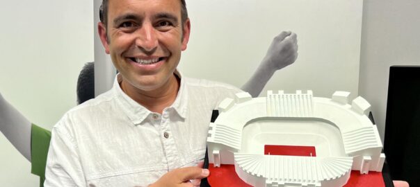 Steven Mifsud, founder of Direct Access, holds a 3D tactile model replica of a football stadium, with braille-enhanced text underneath which reads "Theatre of Dreams".
