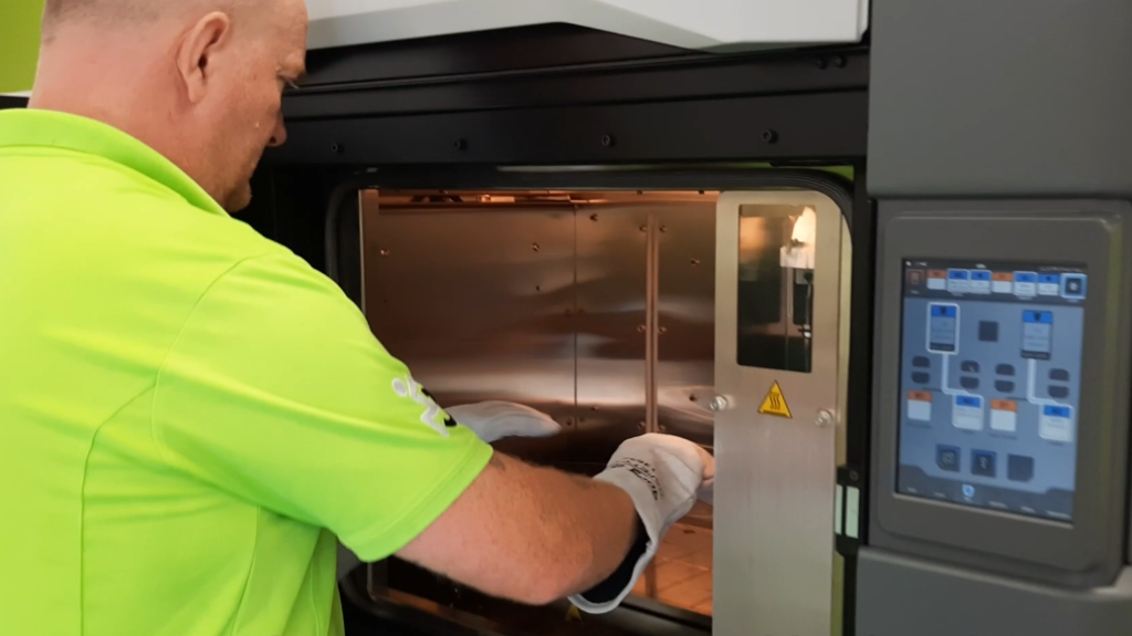 A man in a green polo shirt wearing gloves reaches into a large 3D printer to ready the space for construction of a 3D model.