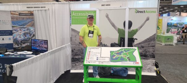 A middle aged man, Andy Lomas, poses for a photo behind a large tactile map board at a product and service conference. In the background is a poster banner for Direct Access showing a young man in a wheelchair cheering on a hillside.