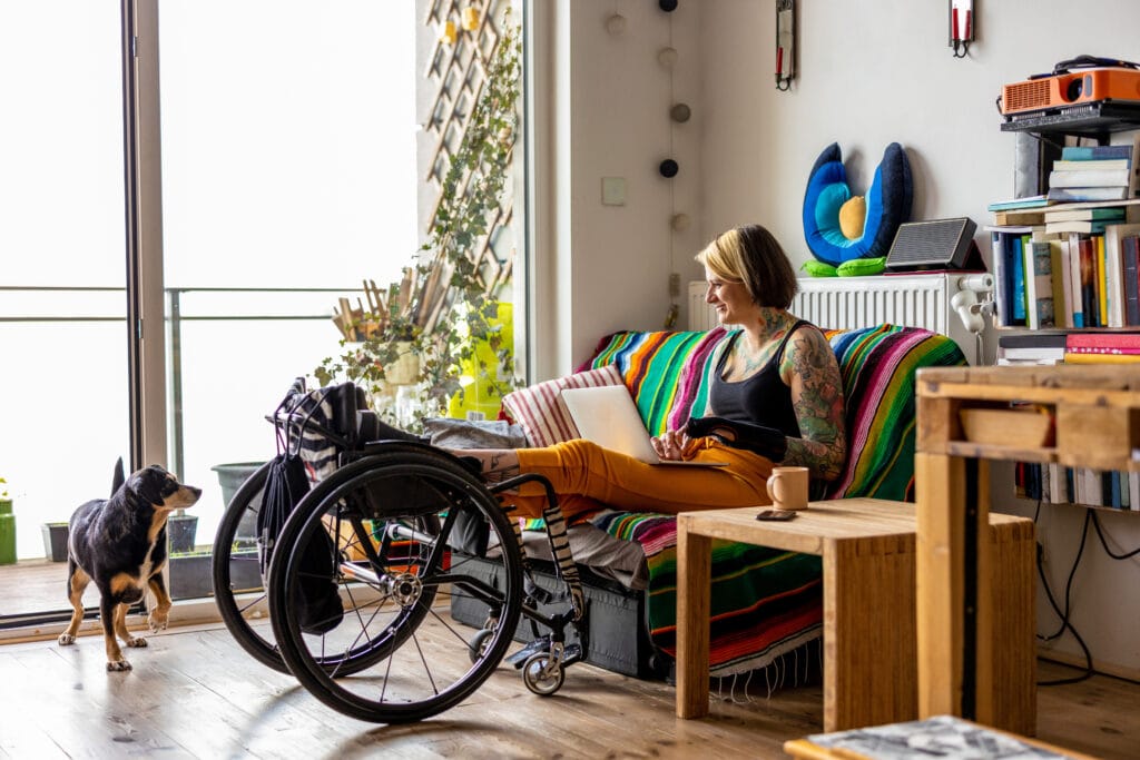 A smiling young woman with a tattoo sleeve on her living room sofa typing on a laptop with her feet held up on a wheelchair. Her brown and black dog watches her.