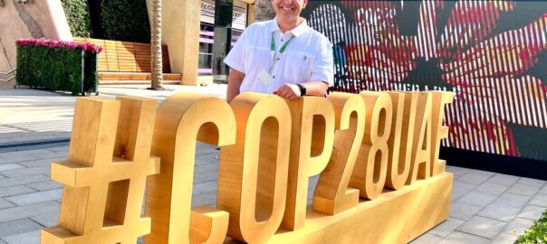 A photograph of a middle aged man wearing a white polo shirt and a hidden disability sunflower lanyard smiles and poses for a picture. His arm is resting on a large structure which reads #COP28UAE