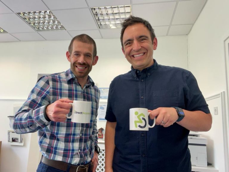 Two male members of the Direct Access team (Steven and Jamie) pose for a photo holding mugs of tea and smiling at the camera in an office space.