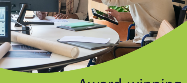 A mixed group of work colleagues discuss work around an office table. One of them, a male wheelchair user, points to his female colleagues monitor to point something out.