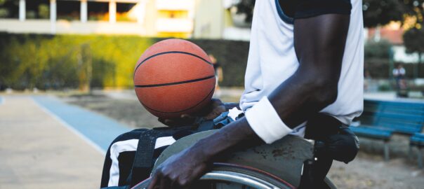 A black wheelchair user wearing a white vest and armband carries a basketball in the palm of his left hand and holds on to his wheelchair with his right. He is on a basketball court on a sunny day.