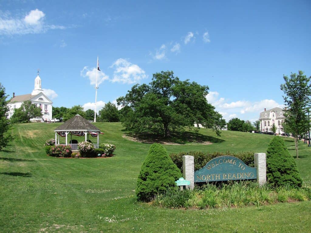 A common with cut green grass and a pavilion surrounded by flowers in the centre. In the forefront is a small garden with a sign which reads "Welcome to North Reading". In the background, a white church and detached house can be seen either side of the horizon.