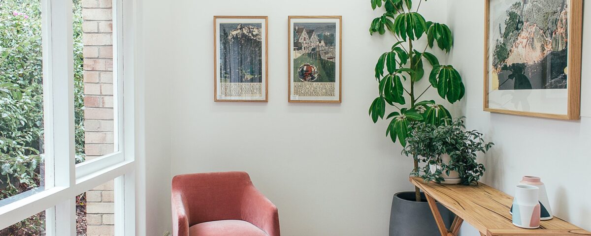 A modern living room space with artificial plants decorating a space which includes a coffee table and red armchairs.