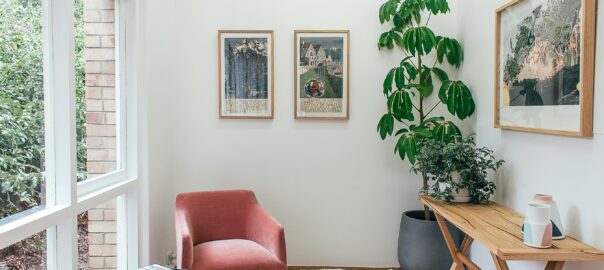 A modern living room space with artificial plants decorating a space which includes a coffee table and red armchairs.