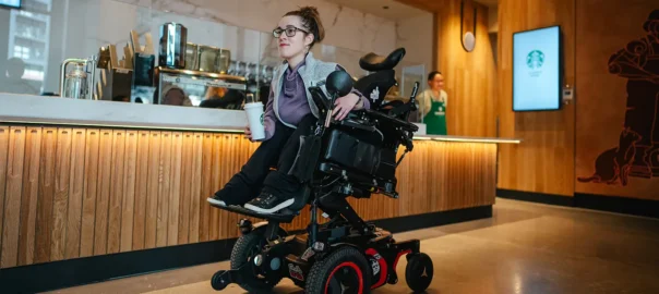 A young Caucasian woman in a height-adjustable wheelchair within a warmly lit coffee shop (the new Starbucks store in Washington). She gazes at something outside the photo. Her hair is tied back and she is wearing glasses, black trousers, and trainers. In her hand is a Starbucks cup.