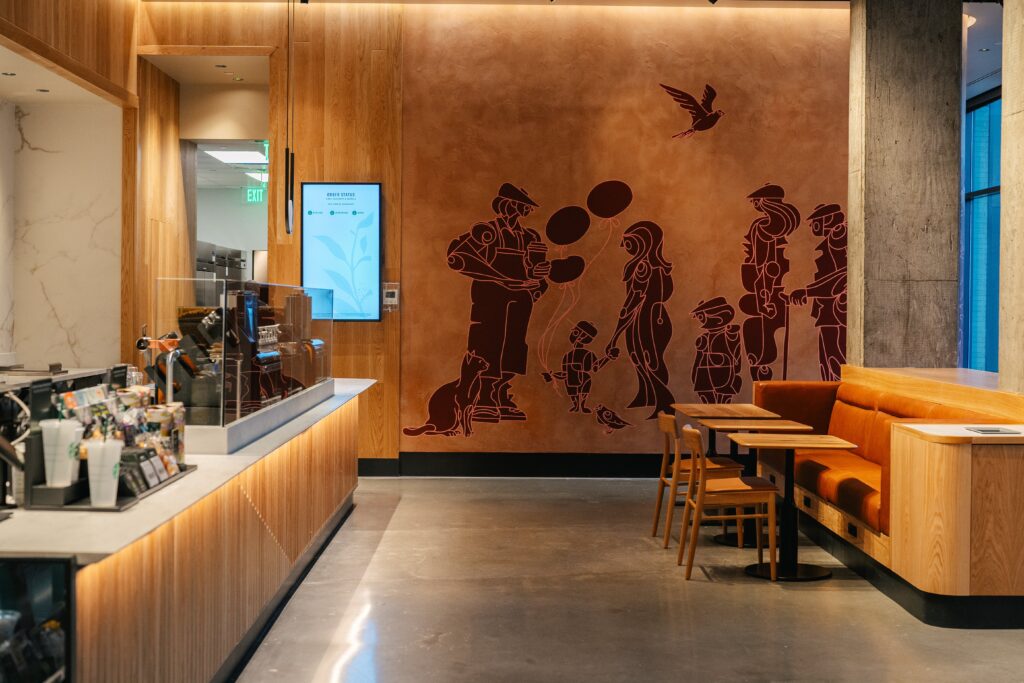 A general view of the warmly lit interior of a new accessible Starbucks store in Washington as seen from front of house. To the left of the image is a counter, to the right is some seating. Central to the image is a mural designed by a deaf artist, showing an abstract, surreal depiction of a man holding a coffee cup alongside a cat, a child holding his mother's hand and a balloon in the other, an older gentleman with a walking stick, a tall woman and a short man. A bird flies over all the characters.