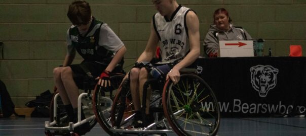 A photograph of two young male and caucasian teenage wheelchair users in dark blue and white sportswear sit on the corner of an indoor basketball court. A bench with a team captain/coach, a middle aged woman, is sat at a table behind them observing the match.