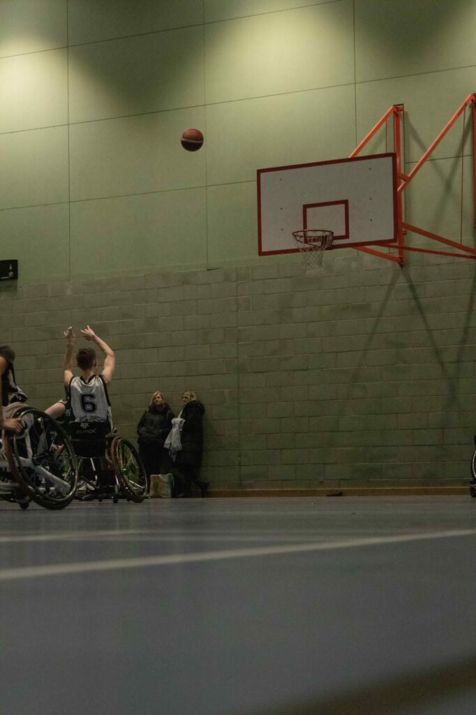 A young male Caucasian teenager in a wheelchair dressed in dark blue and white sportswear on an indoor basketball court. His arms are in the air after throwing a basketball to the hoop. The ball is mid-air.