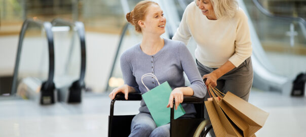 Senior mother with shopping bags laughing and talking to her daughter while pushing her in a wheelchair in a shopping centre.