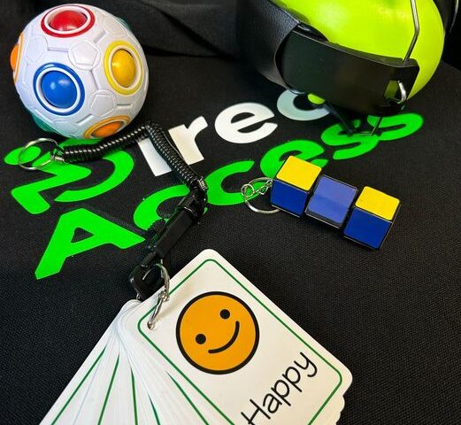 A range of green sensory items including communication cards, headphones and a multi-coloured fidget toy placed on top of a black t-shirt with text that reads Direct Access in white and green.