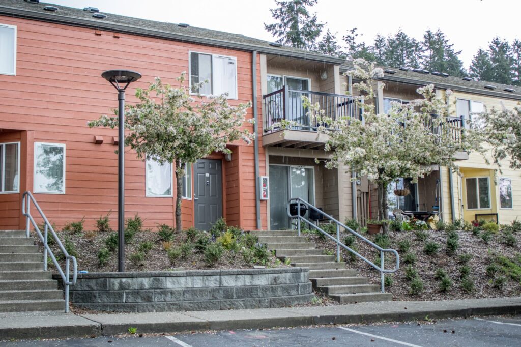 An image of a two-story apartment building in Pierce County, Washington as seen from the parking lot. Several sets of stairs lead to the front of the apartments, with small plants and in bloom trees scattered across the side.