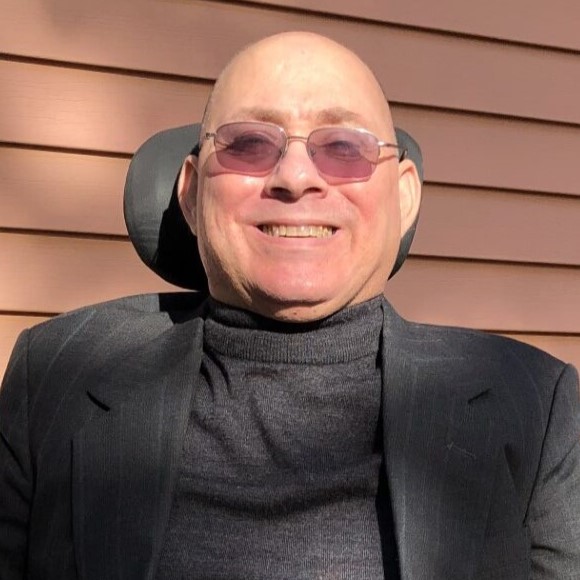 A portrait photo of a bald middle aged Caucasian man wearing a pair of glasses and smiling. He is dressed in a grey neck jumper and grey suit jacket.