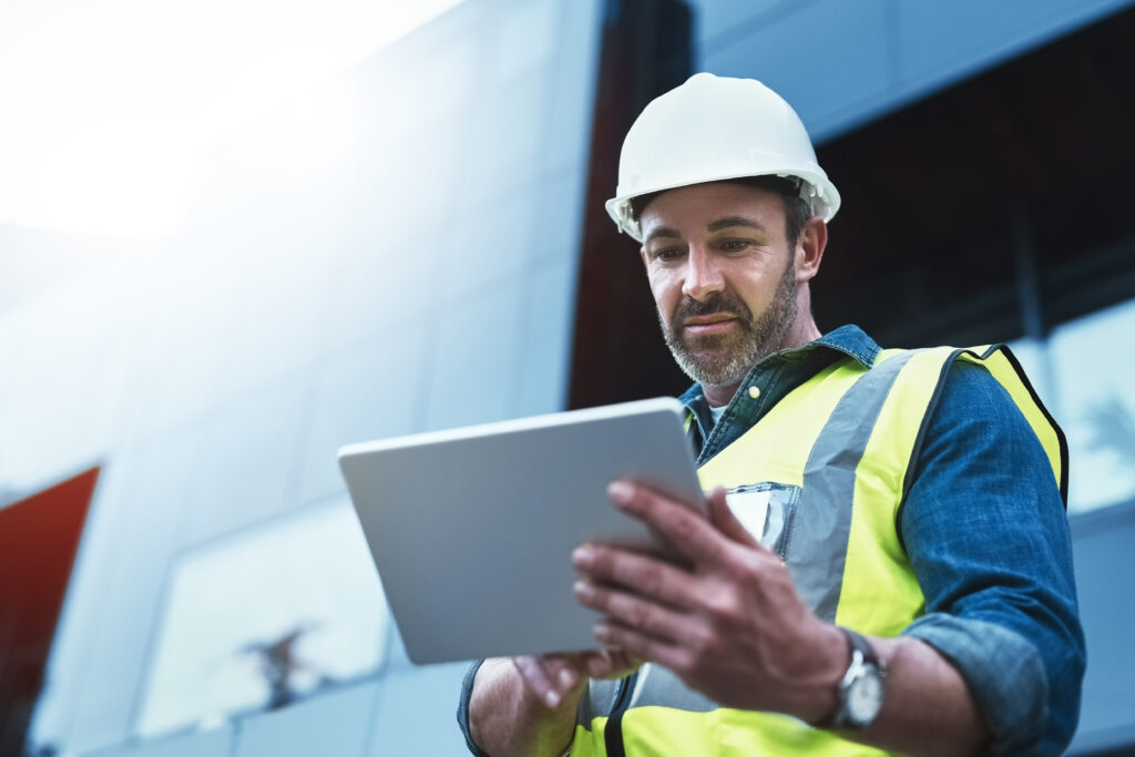 A male engineer on a construction site looks intently on his tablet. Behind him the sun shines and reflects on the surface of a building.