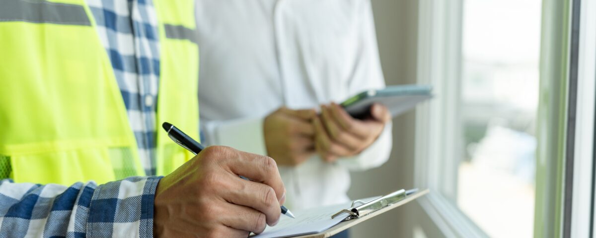 An inspector holding a pen and clipboard writes down something while an inspector holding an electronic tablet from behind observes. Both are stood by a windowsill from within a building.