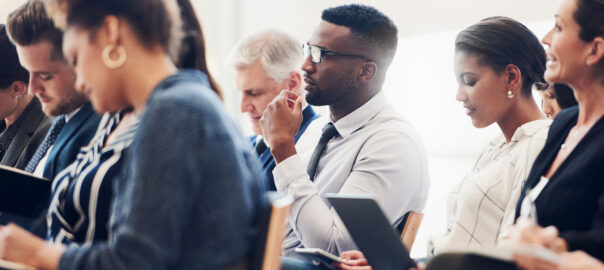 Shot of a mixed group of businesspeople sat on different rows of seats attending a class. Some are watching an out of shot person attentively, while others are writing information on their notebooks, laptops, and tablets.