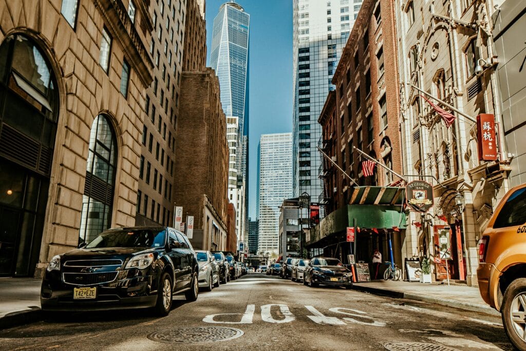 A one-way street in the center of a a non-specific American city. A long row of cars are parked along the side of the road.
