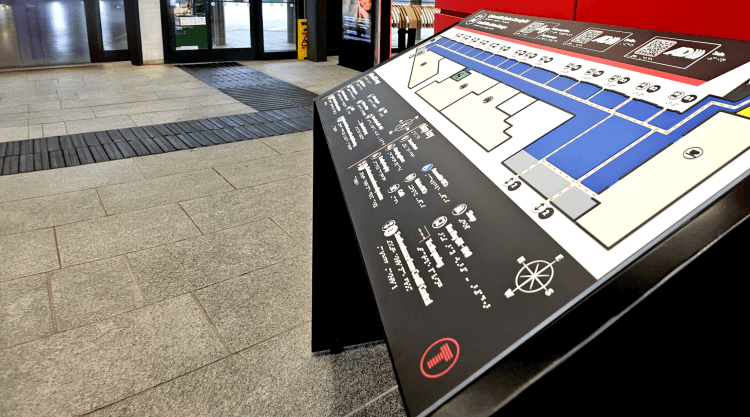 A Direct Access Tactile Map Board as seen from within the Cardiff Bus Interchange. On the map itself is a key, QR codes to scan for audio description and sign language interpretations of the text. The map board is coloured a mixture of black, white, red, and has raised white braille dots embedded into the structure. Directly behind the board is tactile paving leading to the entrance of the interchange.