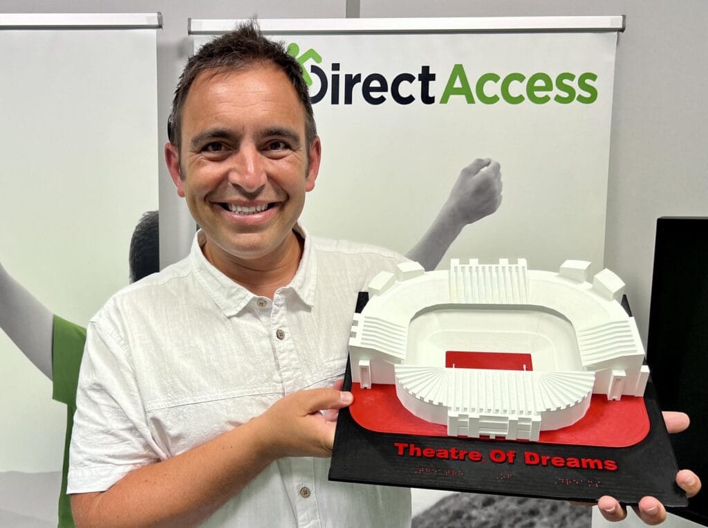 A photo of a middle aged Maltese male, Steven Mifsud MBE, smiling while holding a white tactile model of an arena-like structure called Theatre of Dreams.