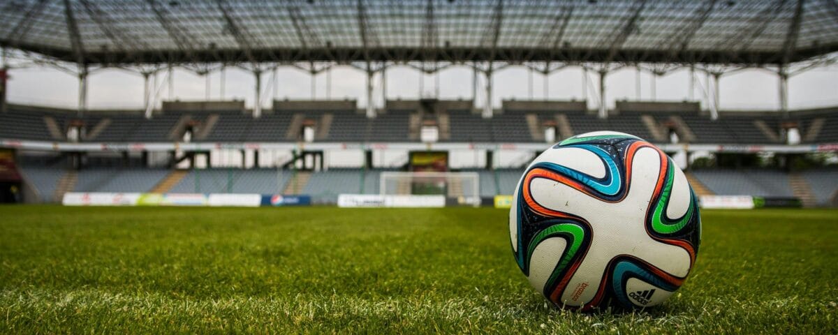 pexels-pixabay-46798 A narrow focus wide shot of a football sat stationery on a stadium football pitch. In the background the stalls of the surrounding stadium and a goal.