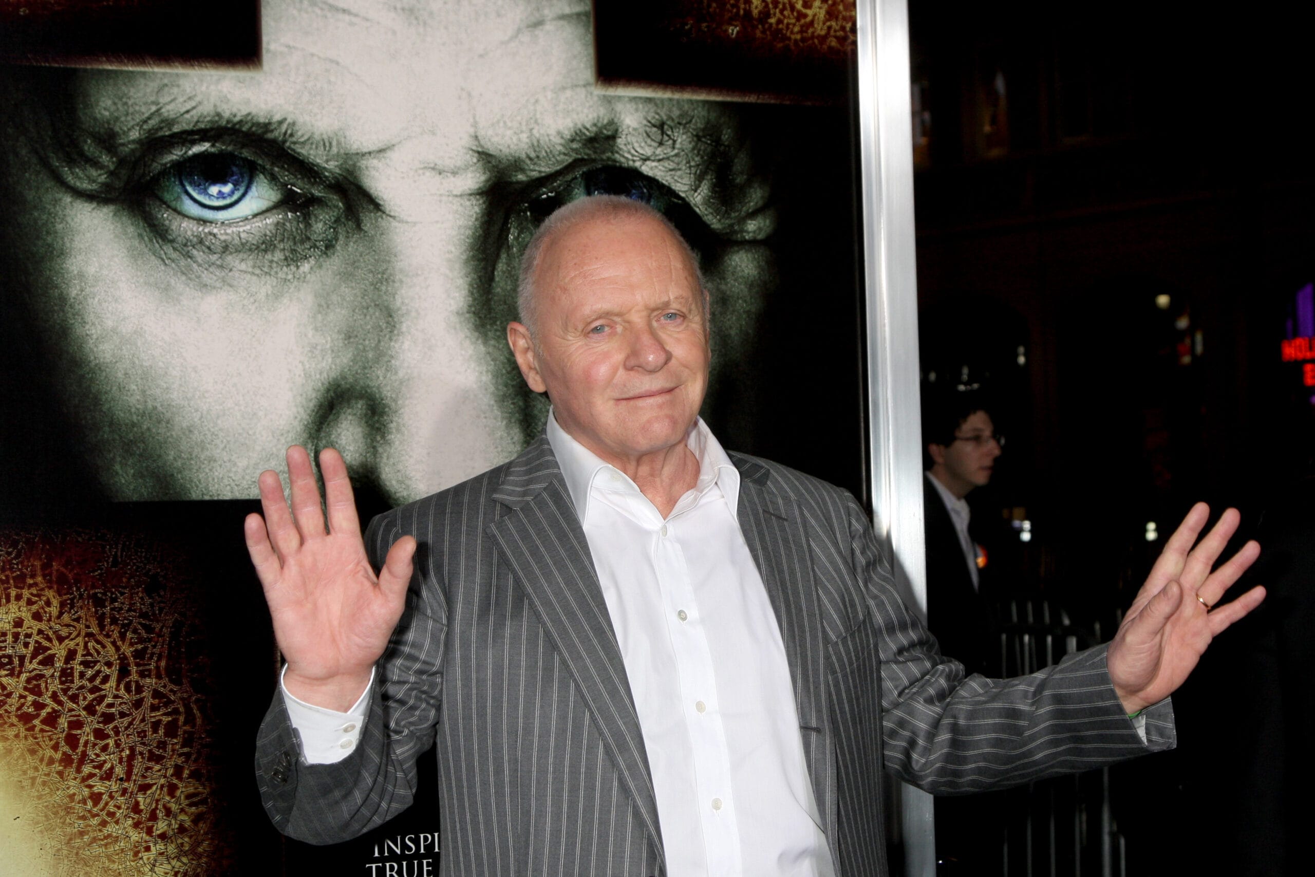 Anthony Hopkins stands in front of a promotional poster for the film The Rite. He wears a gray pinstripe suit with a white dress shirt and raises both hands with open palms, smiling slightly as he poses for photographers. The poster behind him features a close-up of a face with piercing blue eyes and a shadowed cross over the forehead, evoking themes of religion and the supernatural. The setting suggests a movie premiere or press event.