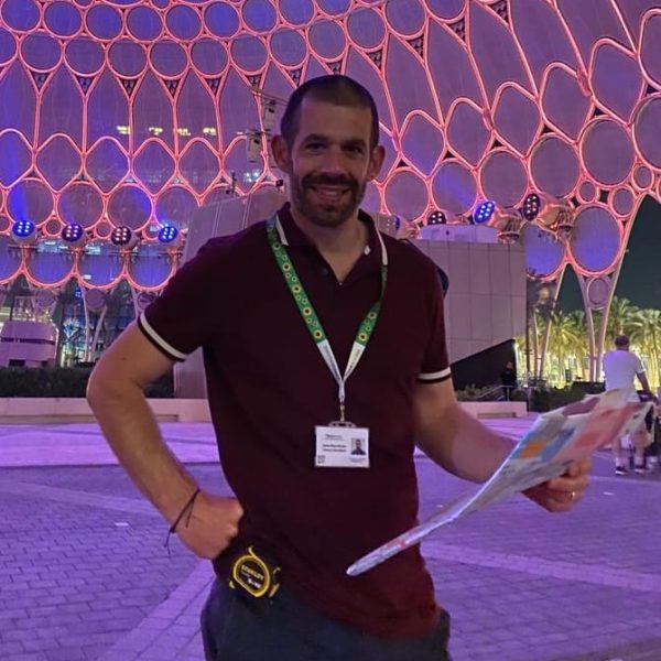 WhatsApp Image 2023-08-23 at 10.49.42 A man holds a large fold-up of a map and poses for a photo in front of a large colourful dome used for night time light shows. He has a tape measure attached to his jeans and is wearing a hidden disability sunflower lanyard.
